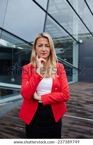 Executive business woman standing against office building looking reflectively to the camera, executive woman carefully listen to the employee, business woman in red jacket standing near office