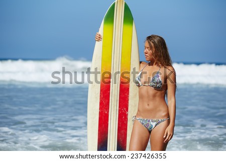 Attractive woman surfer standing with surfboard at ocean seashore, beautiful bikini model on summer vacation engage water sports, at sunny day teenager girl in swimsuit holding her surfboard