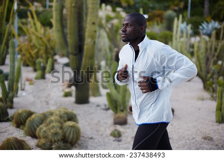 Young fit man in white windbreaker running fast in beautiful park, autumn sport outdoors, attractive dark skinned jogger training outdoors at beautiful evening, sport and healthy living concept
