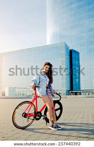 Attractive young woman win fixed gear bike posing outdoors with beautiful skyscraper on background, pretty young brown haired woman standing with her modern pink bicycle in city, stylish hipster girl