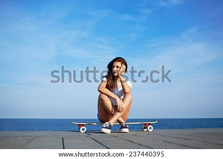 Beautiful sexy girl sits on longboard with amazing horizon of sea on background, young hipster girl posing at beautiful sunset light sitting on her skateboard, attractive woman enjoying sunny evening