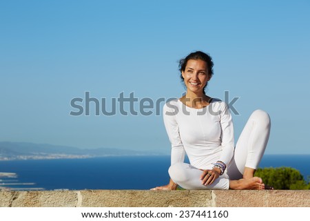 Yoga on high altitude with big city on background, smiling woman seated in yoga pose on amazing city background, woman meditating yoga and enjoying sunny evening, woman makes yoga on mountain hill
