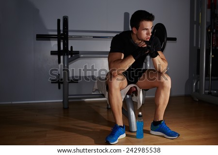 Young attractive athlete sitting on gym equipment with hands against his mouth and resting after fitness training, sad athlete tired to act pensively sitting at gym