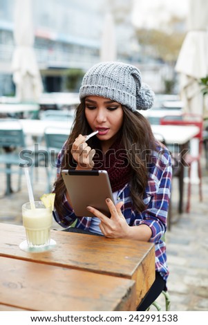 Charming hipster girl rouge lips with pink color sitting in coffee shop, woman applying lipstick outdoors