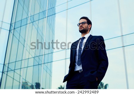 Confident business man in glasses standing against office building with hands in the pockets