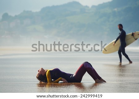 Sexy woman lying on wet suit with beautiful professional surfer on background, young surfer looking to the girl while walk to the ocean