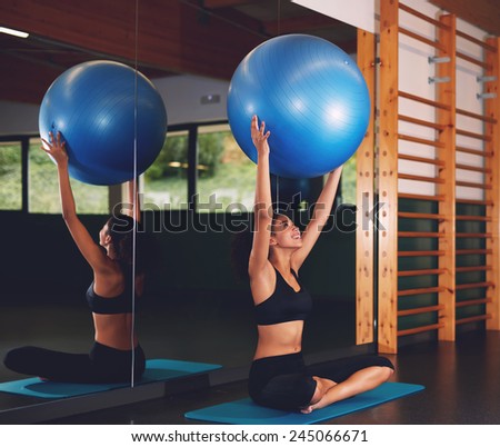 Young athletic woman sitting on the mat with balance ball against big fitness hall mirror