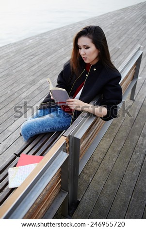 Young asian race woman read the book sitting on wooden bench, charming student girl concentrated reading some book outdoors, stylish tourist girl resting after long walk in new country