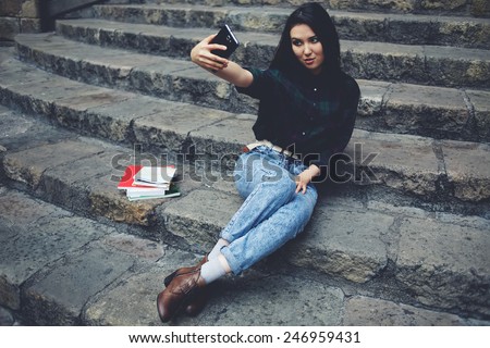 Laughing student girl taking a self portrait with smart phone sitting on steps, beautiful hipster girl photographing herself with mobile phone, charming girl smiling while taking a self-ie outdoors