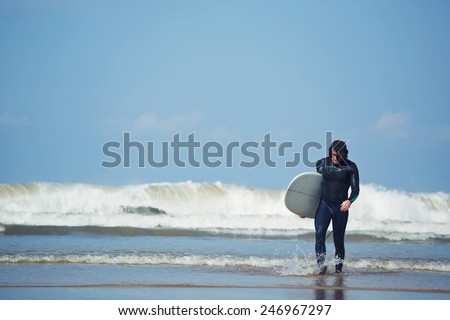 Professional surfer dressed in wetsuit going out of the water after surfing, male surfer going back to the beach holding his surfboard