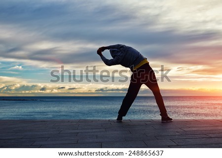 Full length silhouette portrait of a male jogger stretching in the morning and admiring amazing sunrise while standing next to the sea