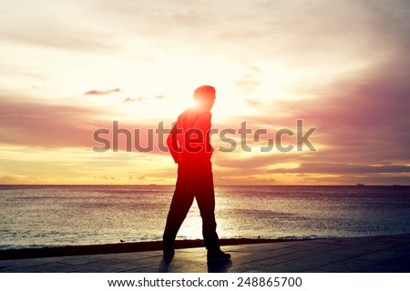 Full length silhouette of a young jogger in sweatshirt relax after workout and admiring beautiful sunrise while standing on the beach