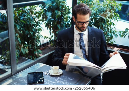 Portrait of a young handsome businessman reading a newspaper at his breakfast in coffee shop, brunette business man holding open newspaper sitting in cafe