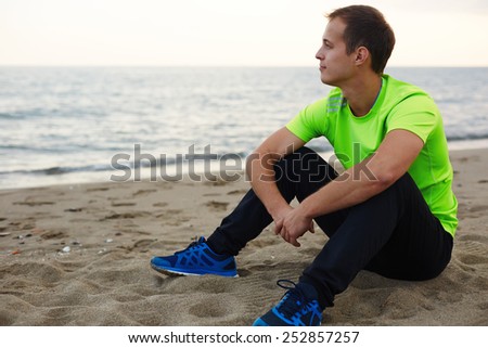 Jogger resting after run sitting on the sand and enjoying amazing sunset view, athlete taking break after training outdoors in amazing landscape, man relaxing outdoors