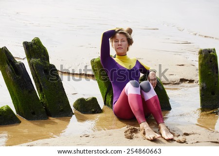 Blonde hair surfer girl relaxing after surf session lying on wooden wreck covered with the sand, summer holidays on the beach