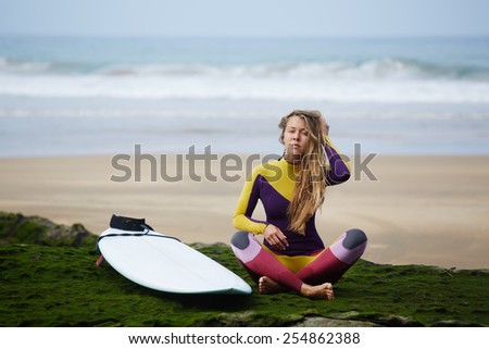 Beautiful professional surfer girl with long blond hair relaxing after surfing sitting against ocean with beautiful waves on background
