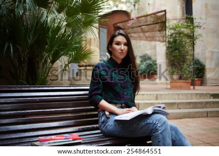 Portrait of nice female student with beautiful brunette long hair sitting with open book on the bench and look to the camera, girl hipster reads book on bench in the summer