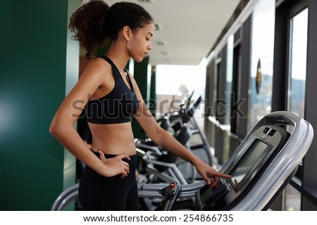 Side view portrait of charming woman with beautiful figure standing on treadmill while touching machine screen, sexy sports woman in bra exercising at gym at beautiful sunny day, focus on the face