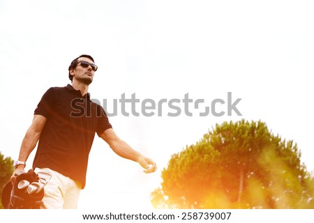 Low angle portrait of professional golf player walking to the next hole holding bag for clubs, good golf game at summer evening weekend on the course, golf player against flare sunset light