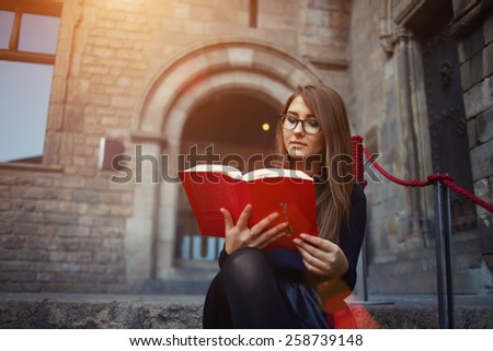 Portrait of a female university student reading interesting book on campus, charming teenager sitting outdoors with open book, attractive young girl read absorbing book at beautiful sunny day, flare