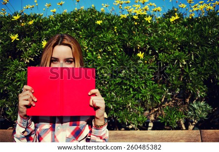 Portrait of charming young woman with pink book held up close to her face, cute female covering half face with a book while standing on spring green hedge with flowers background, filtered image