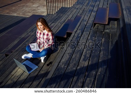 A shot of caucasian college student studying with book and laptop computer at campus, attractive young woman using laptop sitting on wooden staircase enjoying sunny day outdoors, female freelancer