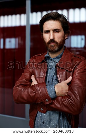 Portrait of fashionable mature hipster man dressed in pattern shirt and leather jacket standing with crossed arms indoors while look to the camera