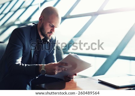 Handsome managing director examining paperwork in bight light office interior sitting next to the window, attractive business man read some documents before meeting, soft focus, filtered image
