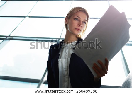 Portrait of young lovely business woman examining paperwork in light office interior sitting next to window, filtered image with flare sun light from the window