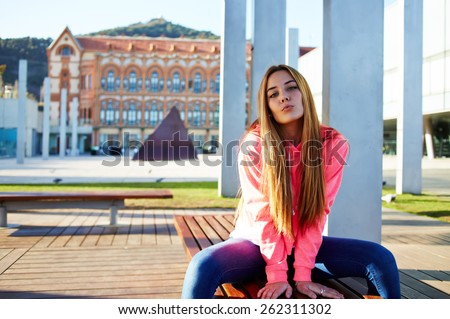 Portrait of attractive school girl posing for camera while giving a kiss, charming caucasian student sitting on the wooden campus bench against university building, cheerful teenager