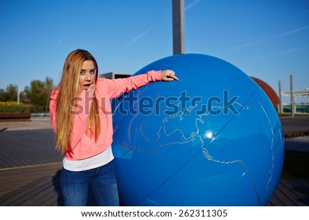 Portrait of young student girl with positive shocking face pointing to the earth globe standing on school campus, science art globe object, female teenager standing near big world globe outdoors