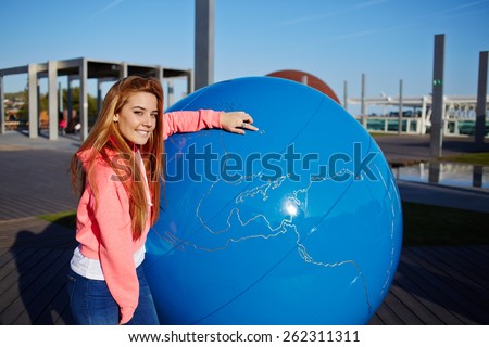 Portrait of smiling student girl pointing to the earth globe standing on school campus, science art globe object, attractive female teenager standing near big world globe outdoors