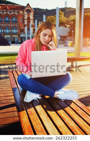 Gorgeous blonde hair female student focused and busy using laptop computer at campus, charming female teenager sitting on park bench with open laptop at spring sunny day, flare sun