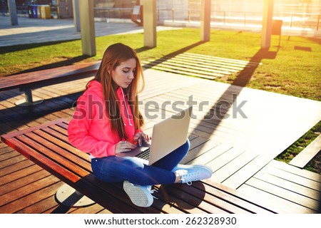 Gorgeous blonde hair female student typing on laptop keyboard sitting at campus, charming female teenager studying for exam on park bench with open net-book and big copy space for text, flare sunshine