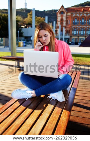 Gorgeous blonde hair female student focused and busy using laptop computer at campus, charming female teenager sitting on park bench with open laptop at spring sunny day, exam preparation