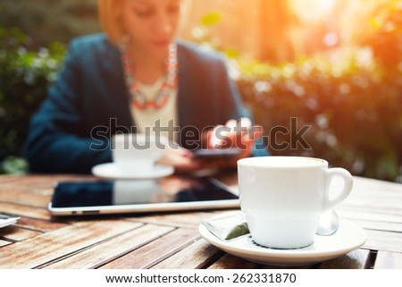 Cup of coffee on the foreground with elegant young woman using busy touch screen tablet at the coffee shop wooden table, work break of business people, flare sun light