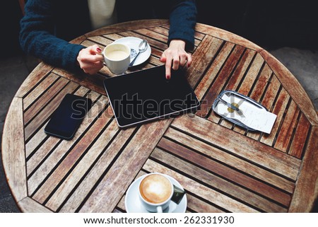 Top shot of a coffee shop table with elegant woman sitting and using touch screen tablet while drink cappuccino, bill check and mobile phone near
