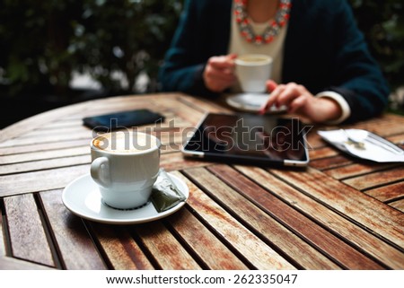 Cup of coffee on the foreground with elegant woman using busy digital tablet at wooden table in a coffee shop