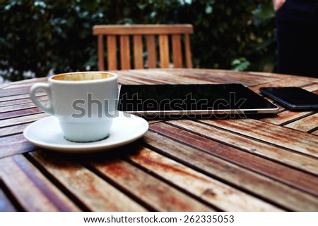 Shot of a digital tablet, smart phone and coffee mug on a wooden table with empty chair, black touch screen computer on cafe table with coffee cup