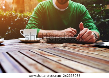 Front view young man sitting at the table with cup of coffee, digital tablet and smart phone, man\'s hands typing message on touch screen tablet while sitting on balcony terrace with plants, flare