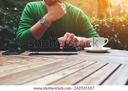 Cropped image male freelancer sitting at the table with cup of coffee, digital tablet and smart phone, man\'s hands typing message on touch screen tablet while sitting on balcony terrace, flare sun