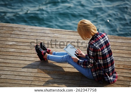 Side view of blonde hair woman using digital tablet while sitting on a wooden pier next to the sea female tourist busy use tablet while sitting outdoors at sunny day girl browsing outside Side view of blonde hair woman using digital tablet while sitting on a wooden pier next to the sea female tourist busy use tablet while sitting outdoors at sunny day girl browsing outside