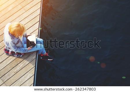 Top view shot of a young blonde hair woman sitting on a jetty next to a sea while using busy digital tablet with a blank screen, tourist woman searching information on tablet while relaxing on a pier