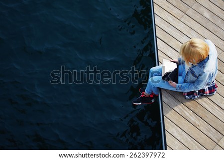 Top view shot of a young blonde hair woman sitting on a jetty next to a sea while using busy digital tablet with a blank screen, tourist woman searching information on tablet while relaxing on a pier