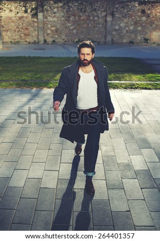 Portrait of elegant fashionable adult man dressed in coat walking in urban setting, stylish hipster man walking on the street at sunny evening, filter