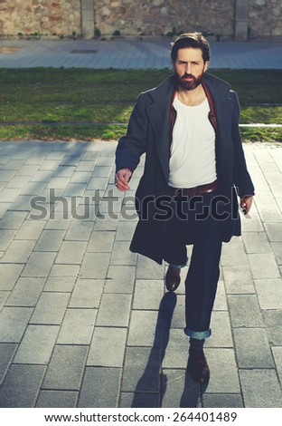 Portrait of elegant fashionable adult man dressed in coat walking in urban setting, stylish hipster man walking on the street at sunny evening, filter