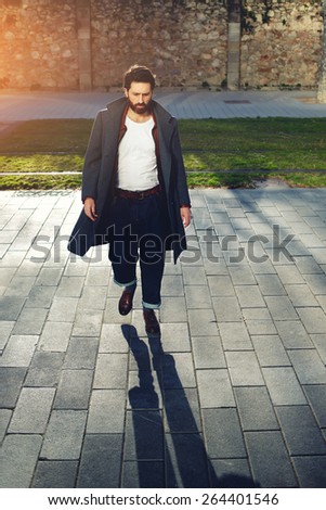 Portrait of elegant fashionable adult man dressed in coat walking in urban setting, stylish hipster man walking on the street at sunny evening, flare sunshine