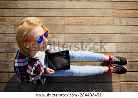 Top view of blonde hair woman in summer colorful glasses using digital tablet while sitting on a wooden pier, female tourist holding tablet while sitting outdoors at sunny day, girl browsing outside