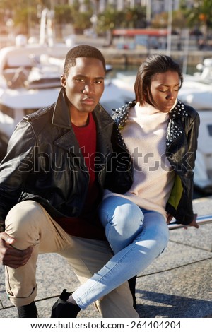 Portrait young fashionable professional models posing outdoors, stylish black couple sitting together in beautiful yacht port of Barcelona