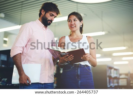 Portrait of young couple of students holding some books while preparing for university exams, business colleagues having fun while standing in modern work space, classmates studying in library, flare